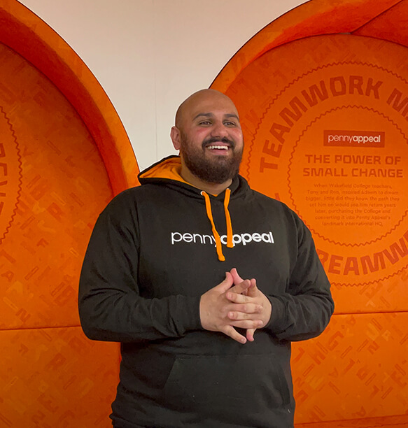 A man standing with a wide smile, wearing a black Penny Appeal hoodie. He is posing in front of a bright orange wall with the Penny Appeal branding and a quote about teamwork.