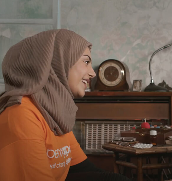A woman with a tan hijab wearing an orange Penny Appeal shirt, seated and engaging in a conversation, with vintage decor and a clock visible in the background.