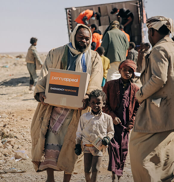 A man holding a 'Penny Appeal Emergency Response' box, with children walking beside him. Volunteers can be seen unloading boxes in the background, all in a dry and barren landscape.