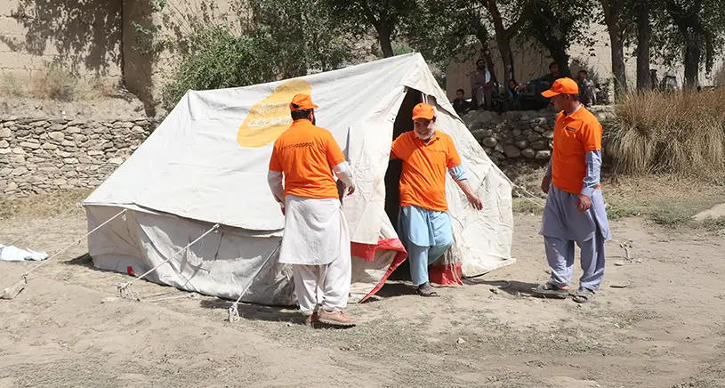 A group of men in Penny Appeal orange clothing stand around a white emergency shelter tent.