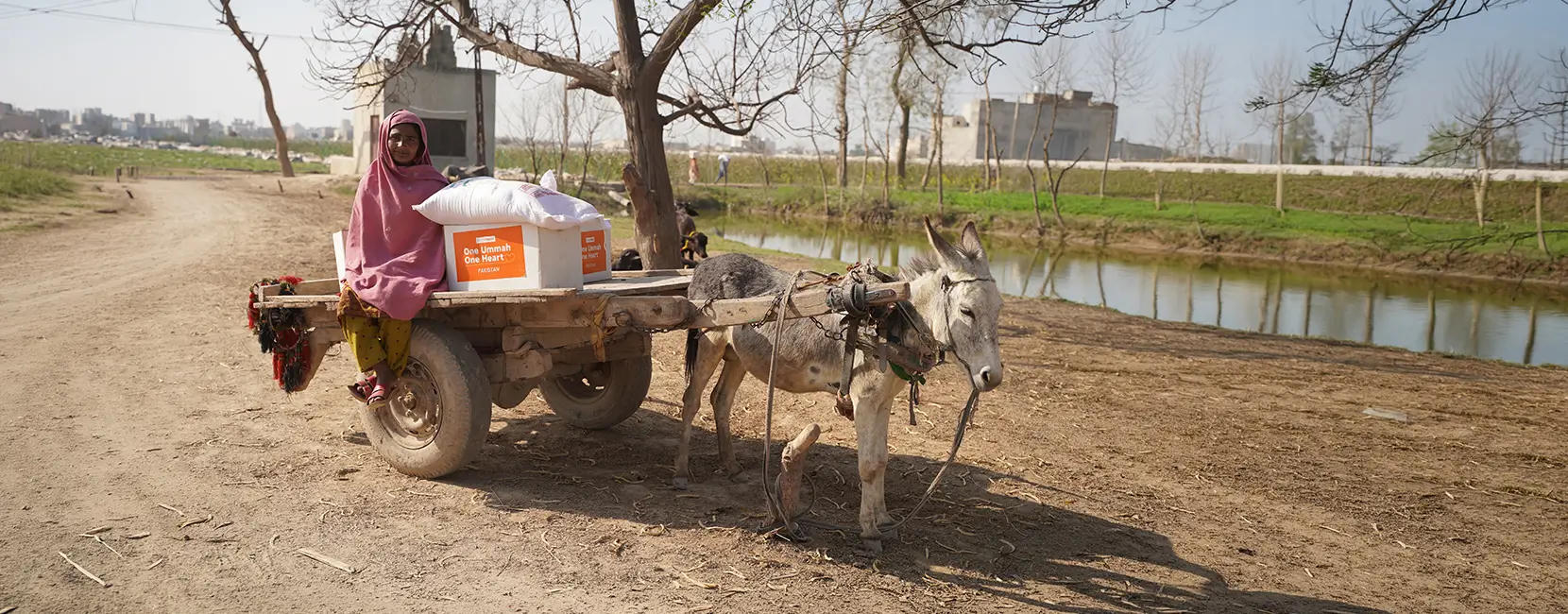 A woman sits on a cart being pulled by a donkey, with a box of supplies from Penny Appeal.