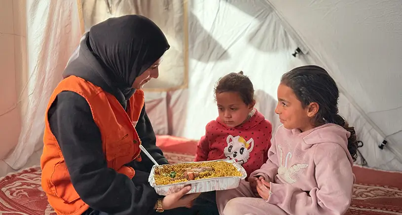 A Penny Appeal worker offers food to two children sitting inside of a tent.