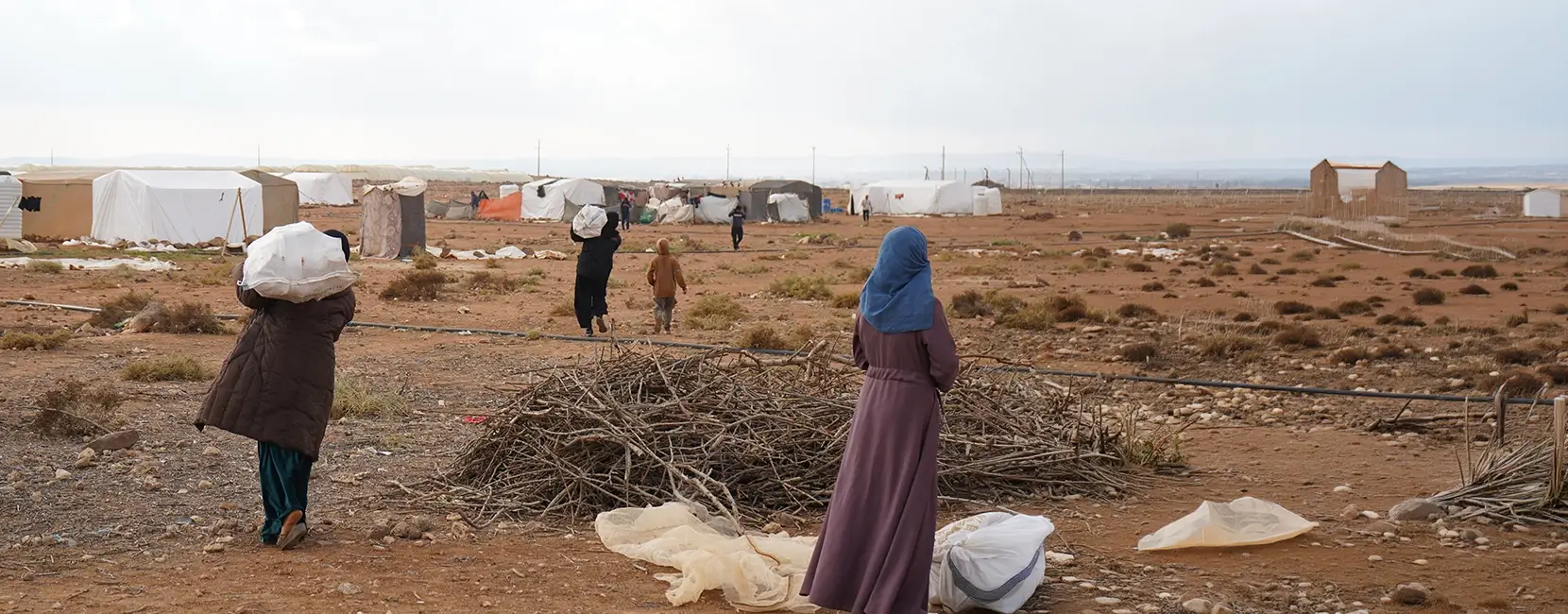 People walking through a refugee camp in Lebanon carrying aid supplies, with tents spread across a dry landscape.