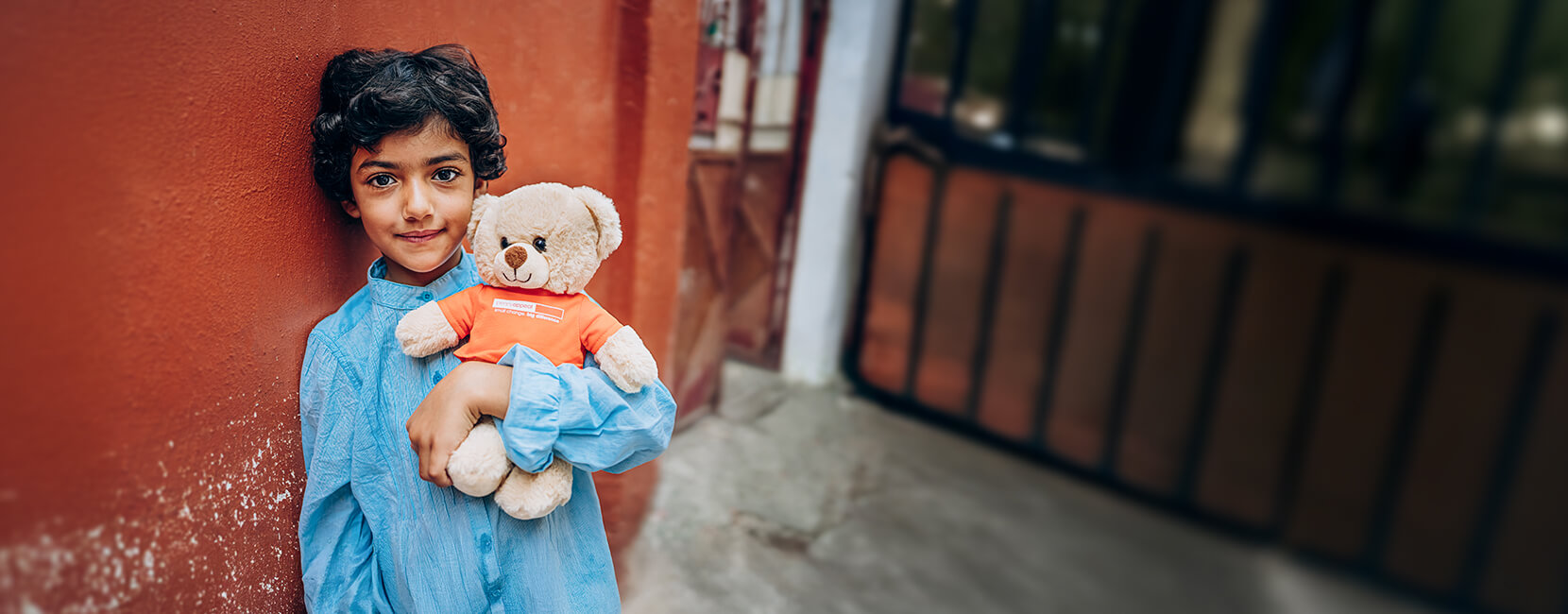 A young child smiling while holding a teddy bear which is wearing a Penny Appeal t-shirt, standing against a red wall.
