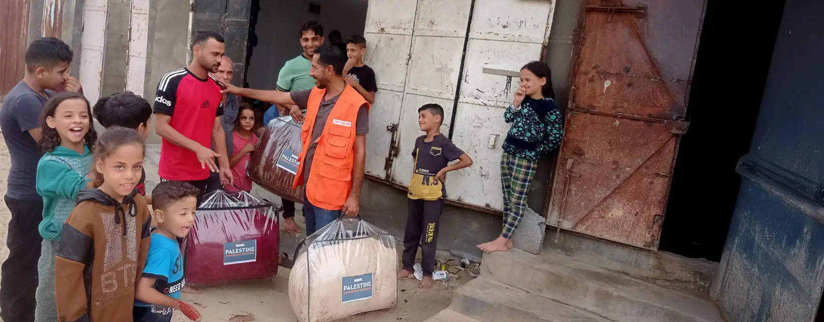 The image shows a Penny Appeal aid worker distributing winter blankets labeled "Palestine Winter Aid" to a group of smiling children and adults outside a building.