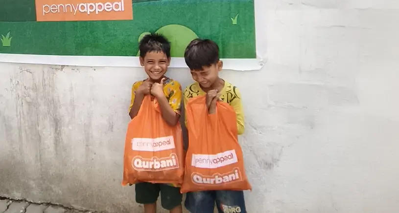 Two children smile at the camera while holding Penny Appeal Qurbani bags.