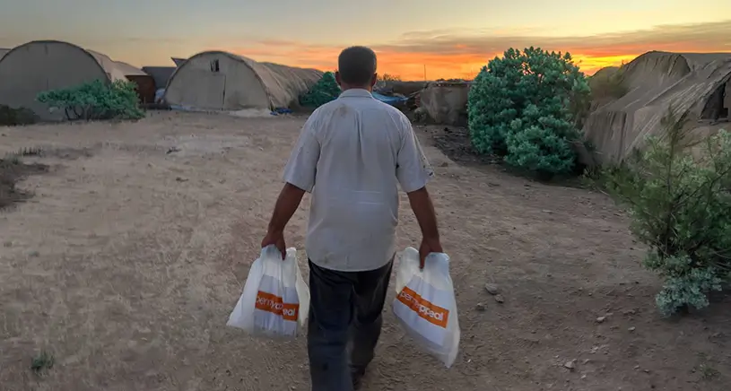 A man carried two bags of meat from Penny Appeal through a refugee camp at sunset.