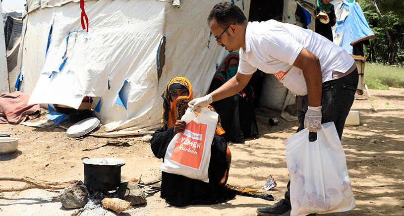 A Penny Appeal aid worker hands a bag of Qurbani meat to a woman sat on the ground.