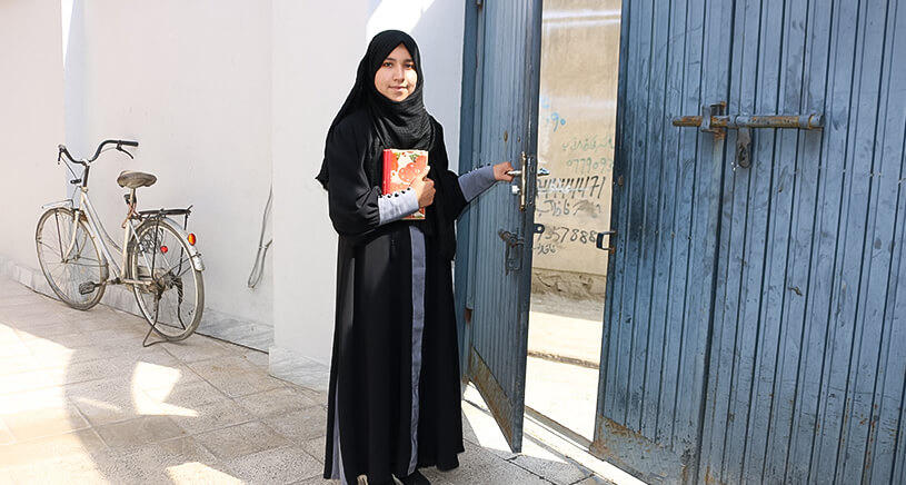 A girl wearing a hijab and all black clothing holds the Quran and smiles at the camera