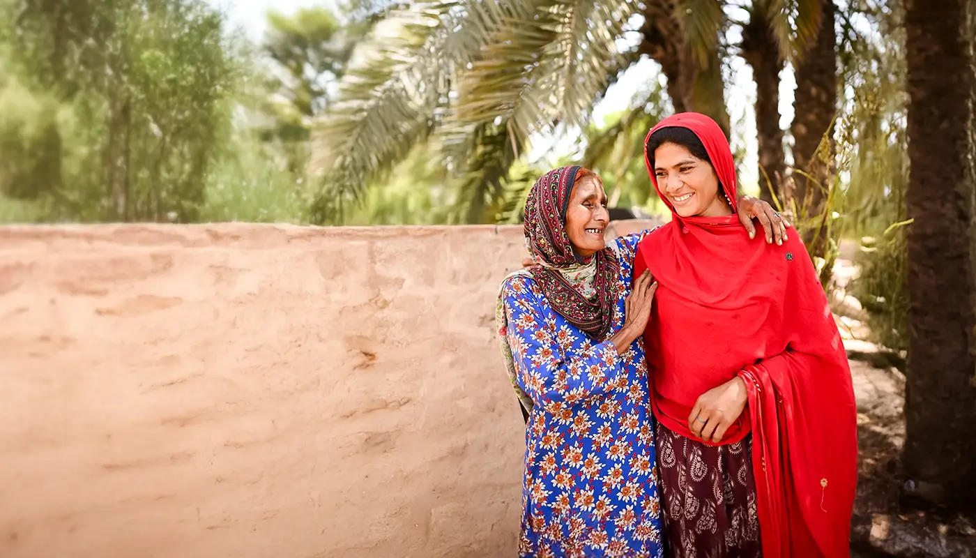 Two women in colorful headscarves smile and embrace outdoors beside a sunlit wall and palm trees.