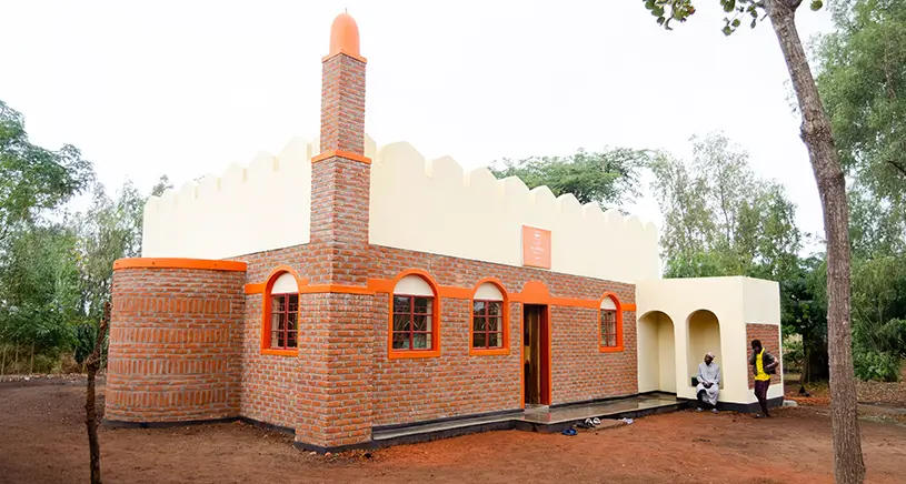 A small brick mosque with orange accents and a cream-colored upper section, featuring a minaret on the left and arched windows and doorways. Two people are sitting near the entrance, which is shaded by an arched porch.