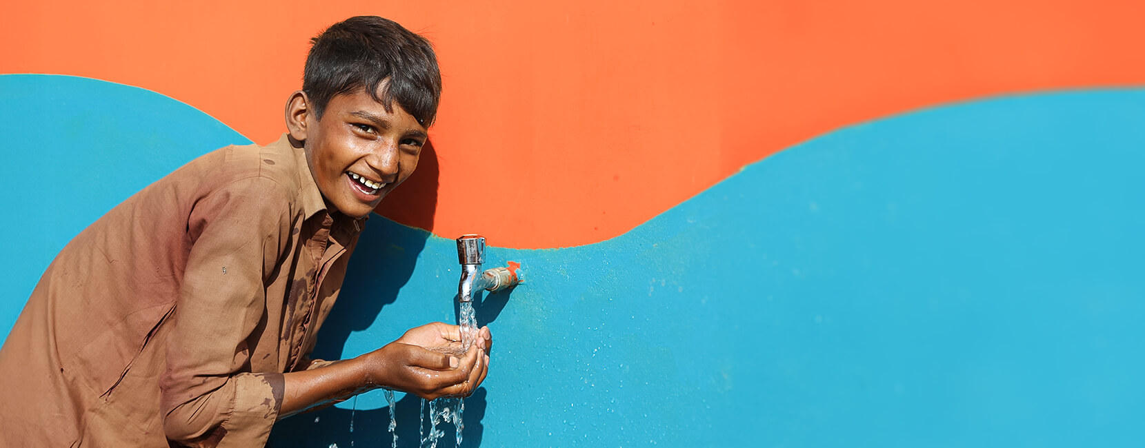The image shows a joyful boy smiling while collecting water from a tap mounted on a brightly painted wall (orange and blue). His hands are cupped to catch the flowing water, and he appears happy and refreshed.