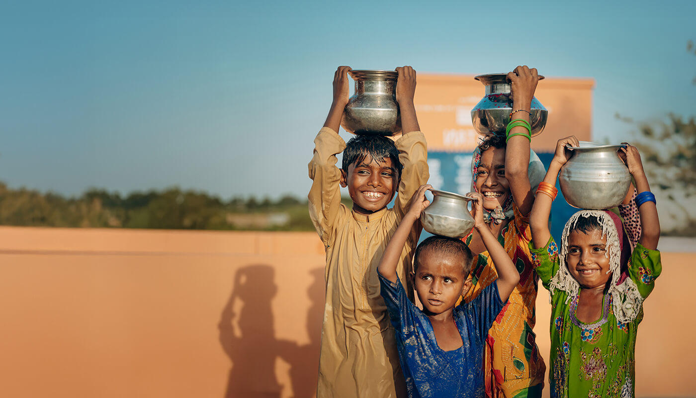 Smiling children hold metal pots on their heads after collecting clean water.