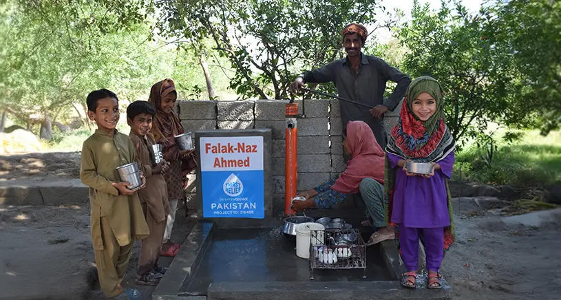Children and a man gather around a hand pump collecting clean water. The pump features logos for Penny Appeal's Thirst Relief campaign. The children smile while holding containers.