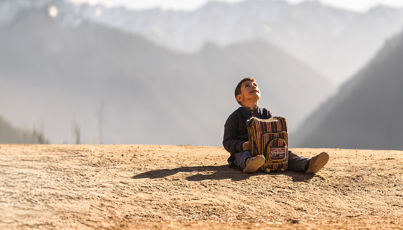 A child with a backpack sits on the ground and looks up at the sky.