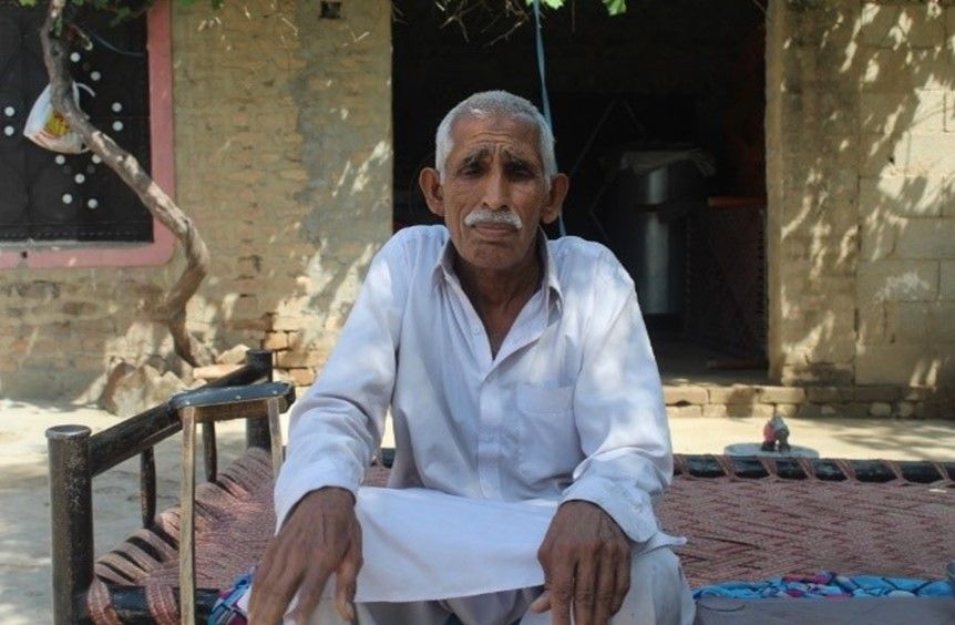 This image shows an elderly man sitting on a traditional woven charpai (cot) outside a modest home with a rustic brick and cement wall. He is dressed in simple, traditional white clothing, common in rural South Asia.