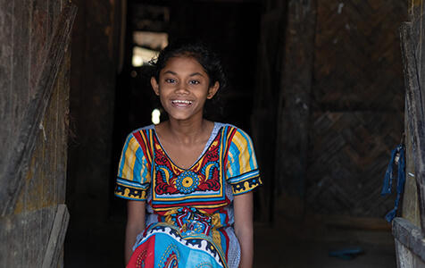 A young girl smiles brightly while sitting on the floor in a doorway. She is wearing a colorful traditional dress, featuring vibrant patterns and intricate designs.