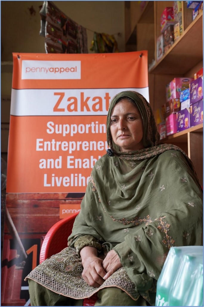 A lady in traditional clothing sits inside of a small shop with a Penny Appeal banner in the background.