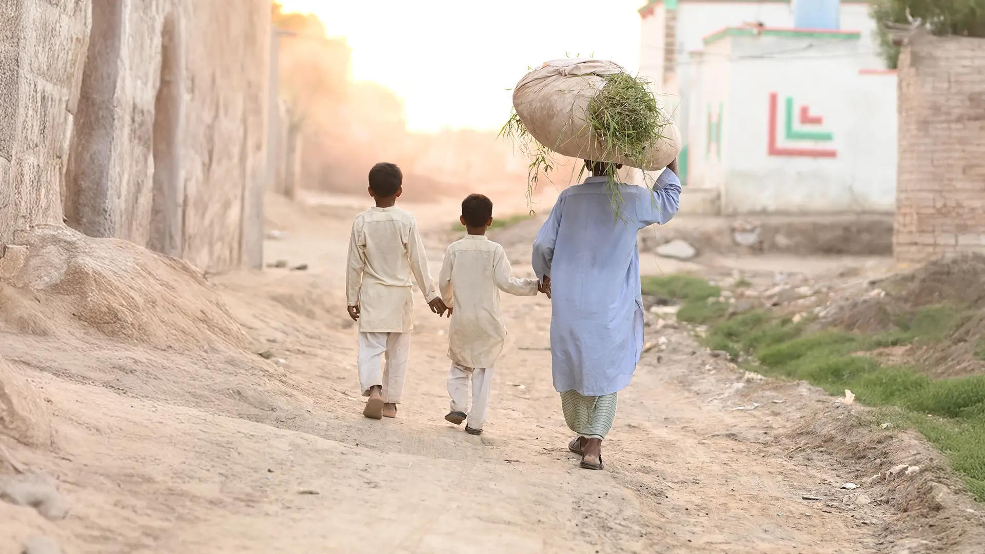 A person wearing traditional South Asian clothing walks down a dusty village path with two children, holding their hands. The person carries a large bundle of grass on their head. Sunlight glows in the background, illuminating simple homes and walls.