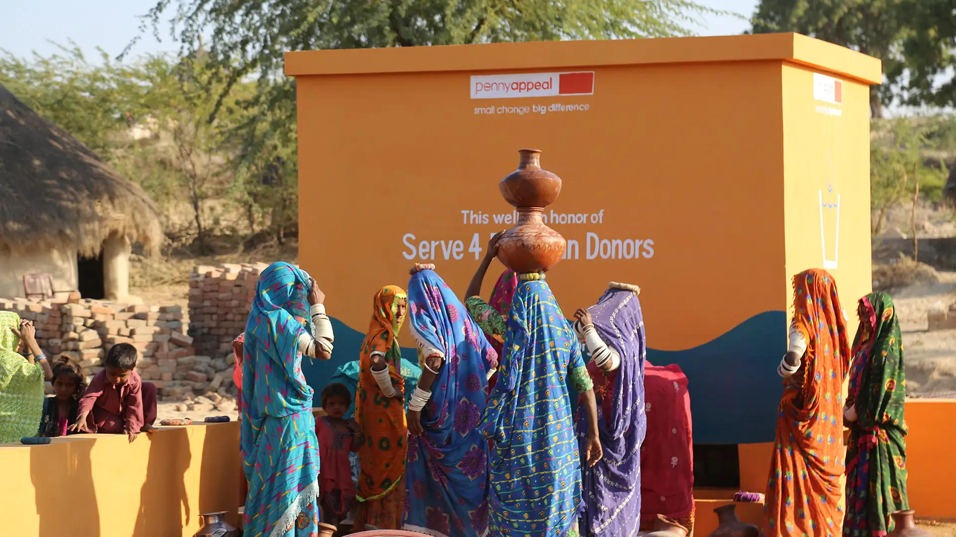 Women in colorful traditional clothing collect water from a Penny Appeal Thirst Relief Deep Well in a rural village, carrying clay pots on their heads near an orange water structure.