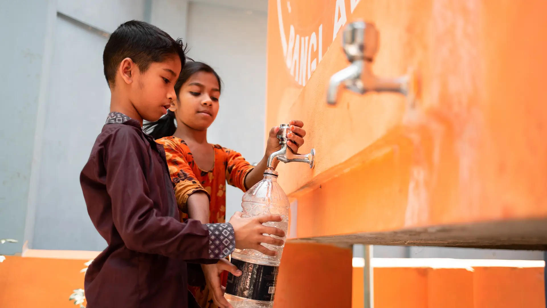 Two children fill a plastic bottle with clean water from a Penny Appeal Thirst Relief Deep Well tap station painted orange.