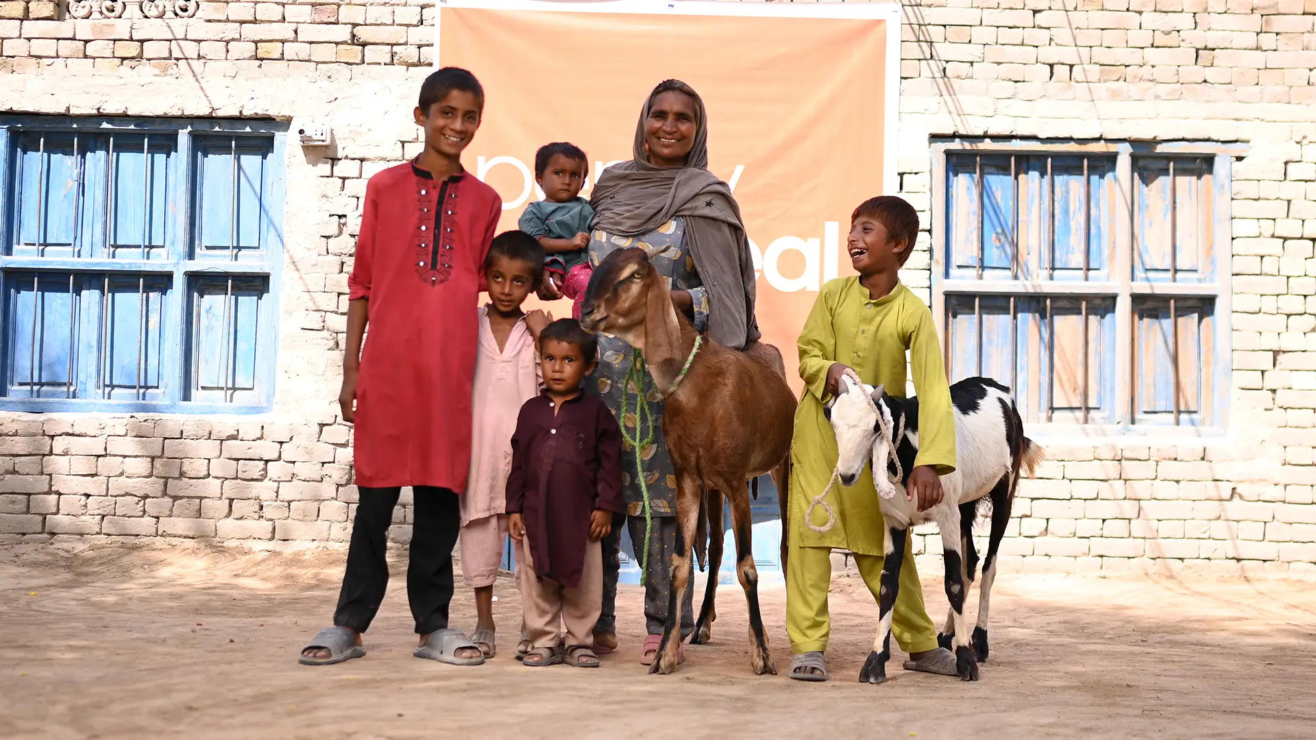 A smiling family stands with their goats outside their home, supported by Penny Appeal.