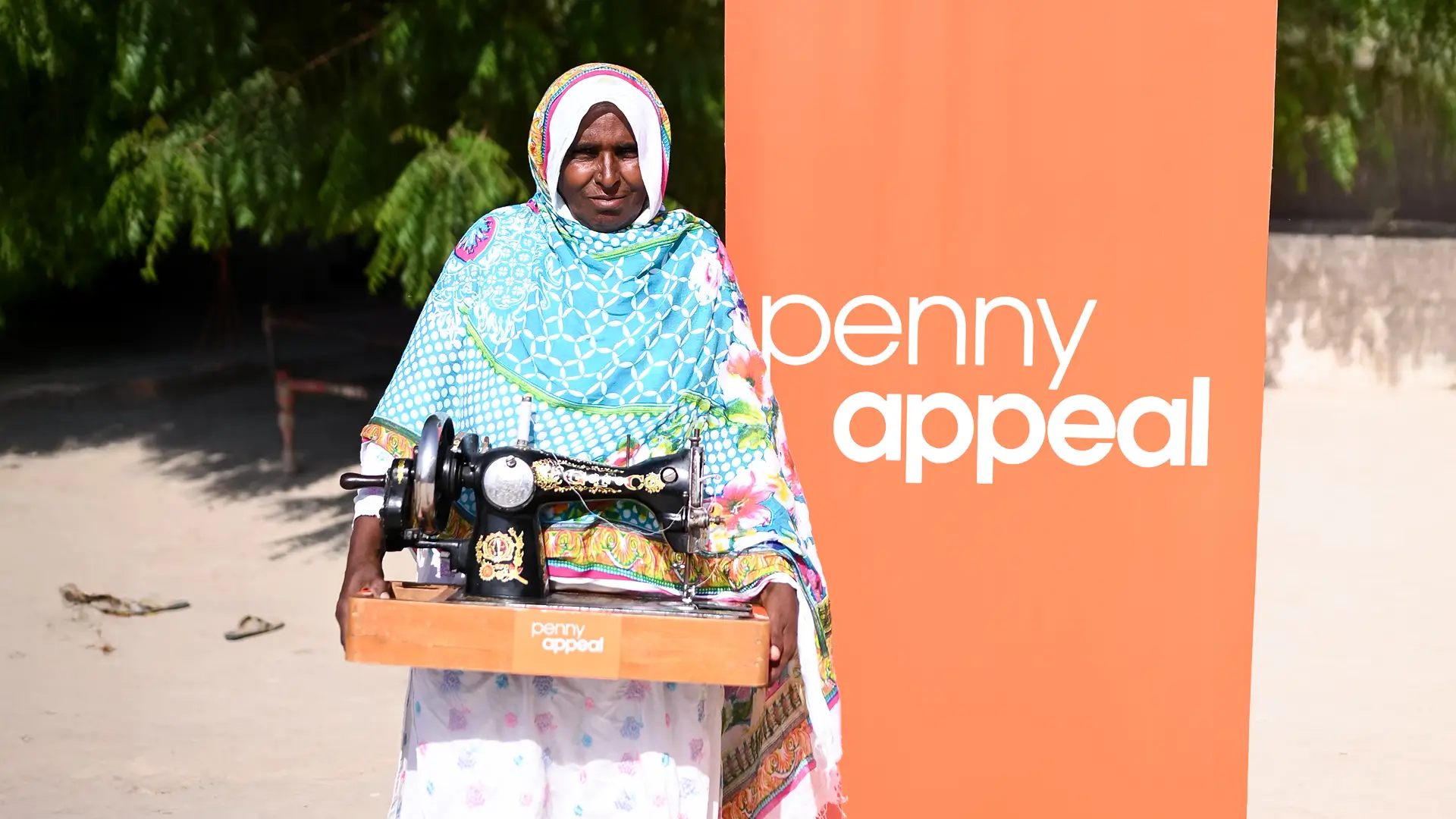 A woman in traditional clothing stands in front of a Penny Appeal sign, holding her new sewing machine.