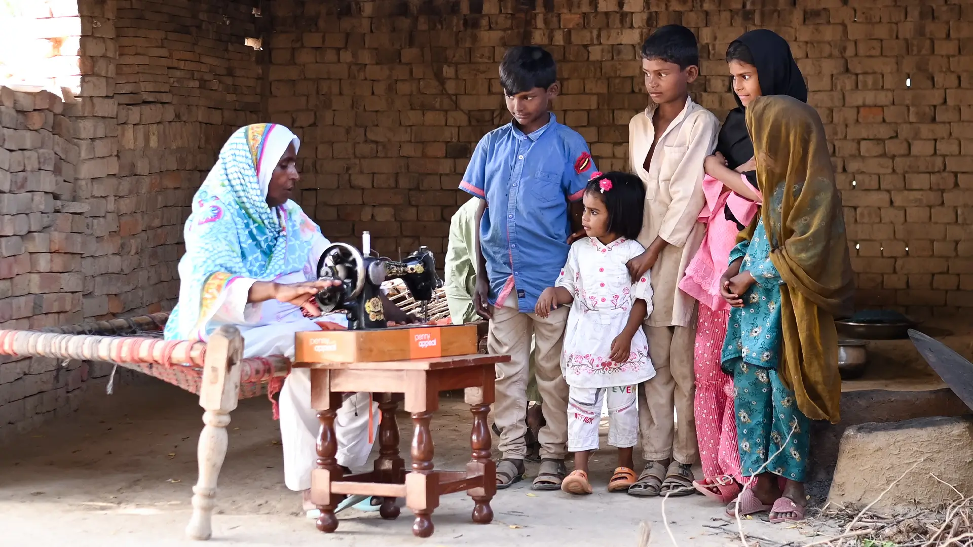 A woman in traditional clothing uses her Penny Appeal gifted sewing machine while a group of children stand and watch.