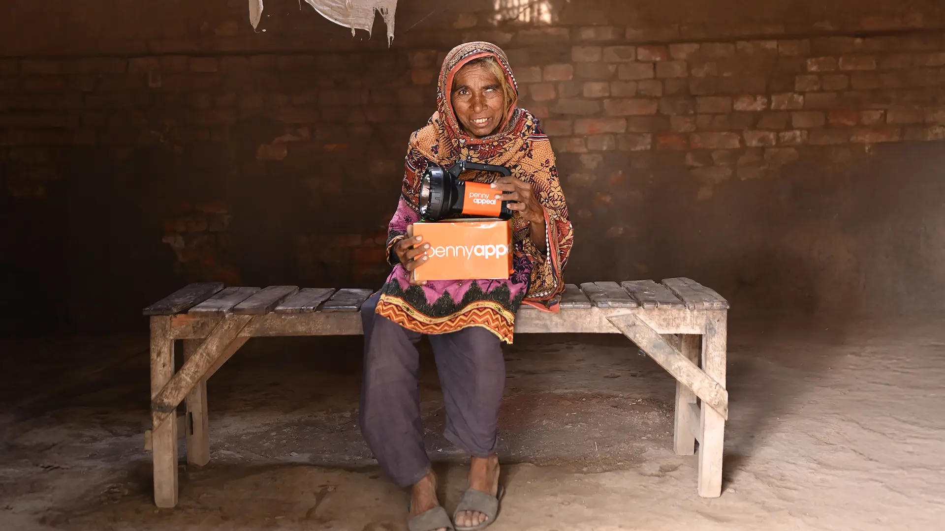 An older woman sits on a wooden bench inside a modest room with brick walls, smiling as she holds a solar lamp and an orange box labeled “Penny Appeal.”.