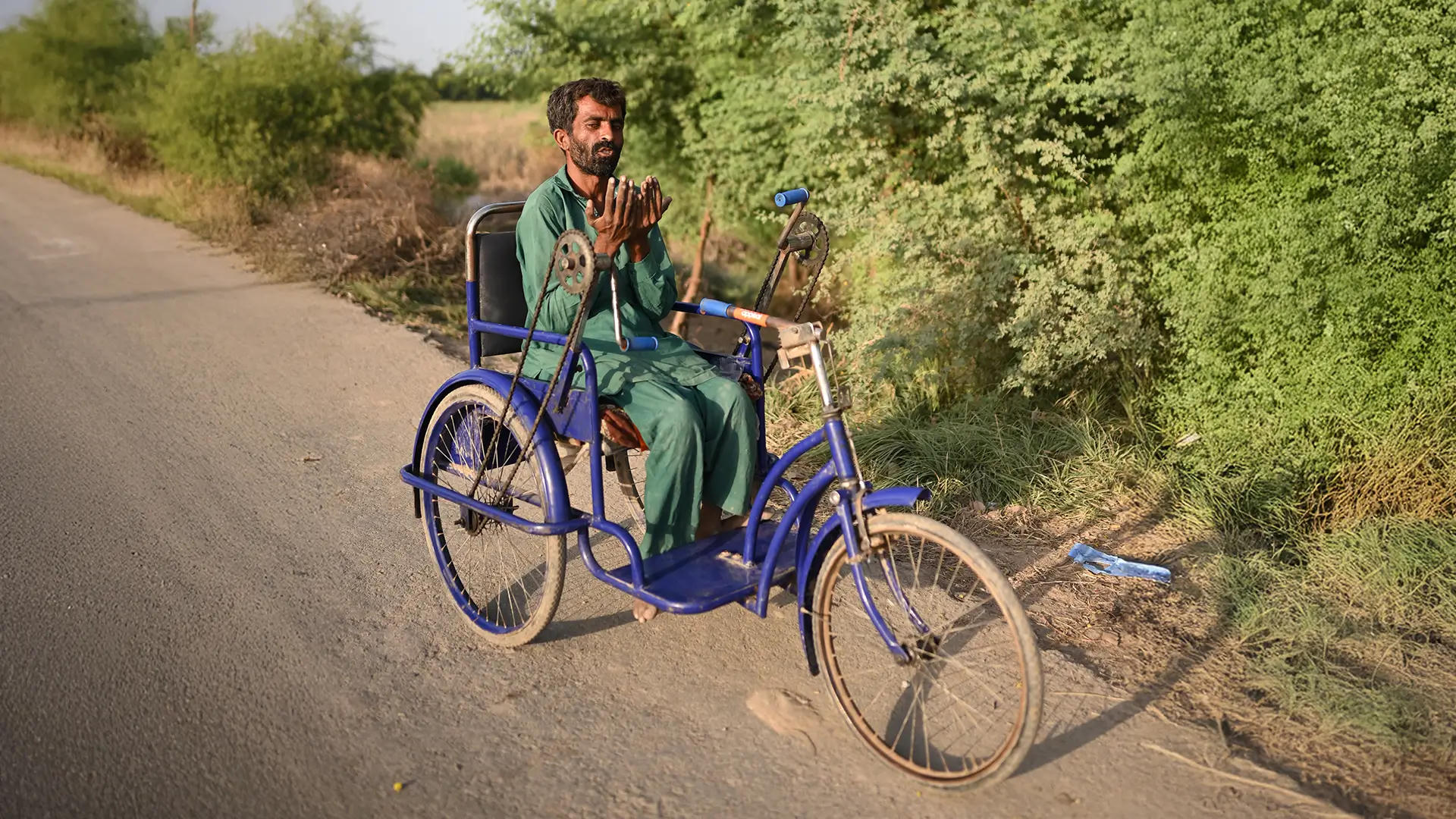 This image shows a person using the specialized wheelchair, designed like a pedal-powered tricycle, in a rural area. The wheelchair is branded with the Penny Appeal logo.