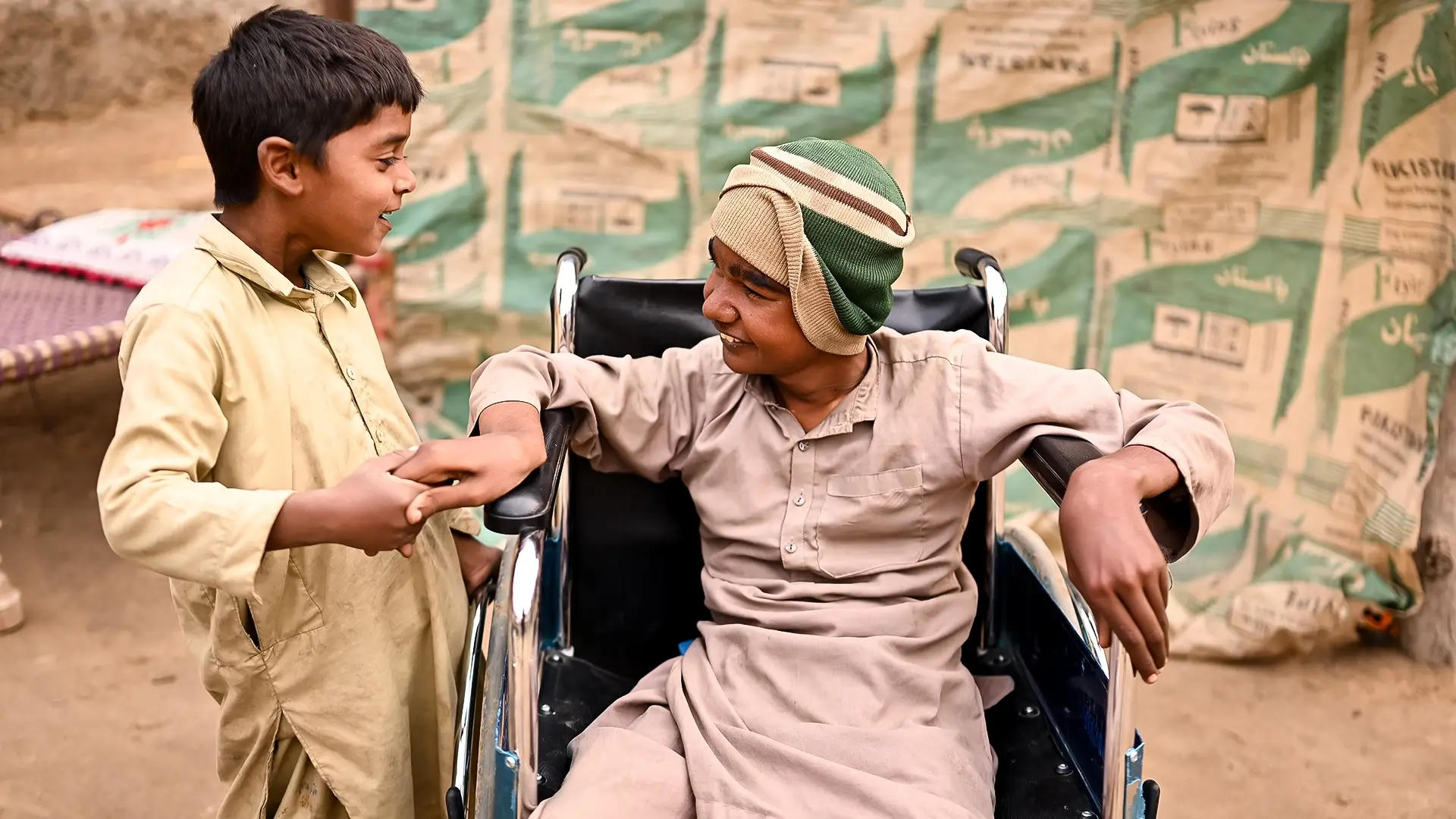 A smiling child sits in a wheelchair and speaks with his friend stood nearby.