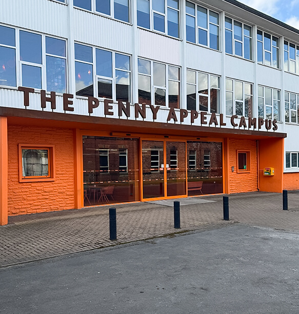 The exterior of The Penny Appeal Campus building. It has large windows and an orange-colored entrance with the words 'THE PENNY APPEAL CAMPUS' prominently displayed on the wall.
