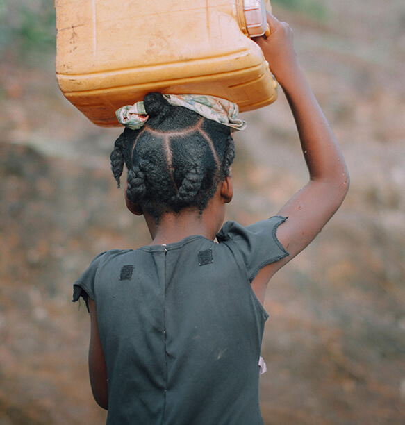 A young girl carrying a large yellow water container on her head. She is wearing a simple black dress, and her hair is tied in small braids with a scarf around her head. The background is a blurred earthy, rocky terrain.