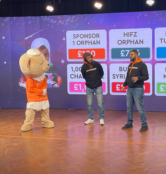 Two men on stage at an event with a mascot dressed in an orange bear costume. The backdrop displays fundraising details for various charitable causes, with the men wearing black Penny Appeal hoodies and speaking to the audience.