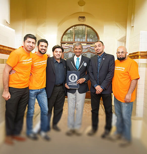 A man holding a 'Penny Appeal Emergency Response' box, with children walking beside him. Volunteers can be seen unloading boxes in the background, all in a dry and barren landscape.
