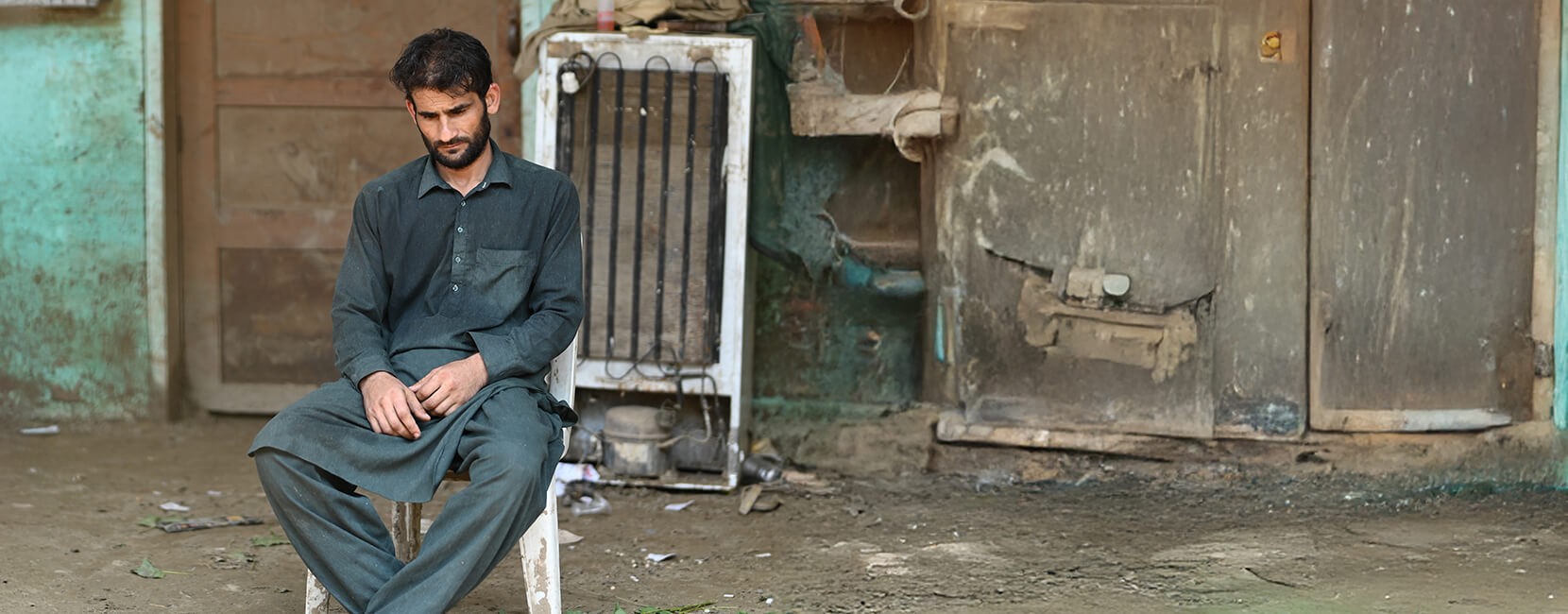 A man in dark traditional clothing sits alone on a plastic chair in front of a weathered building, looking down with a pensive or somber expression. The surrounding area appears dirty and run-down, with scattered debris and old appliances in the background.