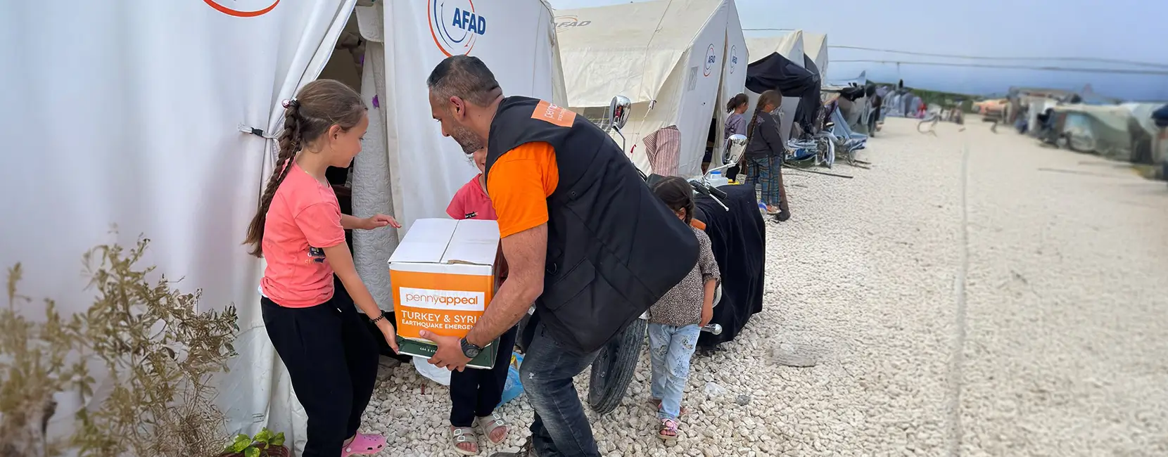 A Penny Appeal aid worker hands a box of emergency supplies to a young girl outside a tent in a refugee camp, as part of the Turkey & Syria Earthquake emergency response.