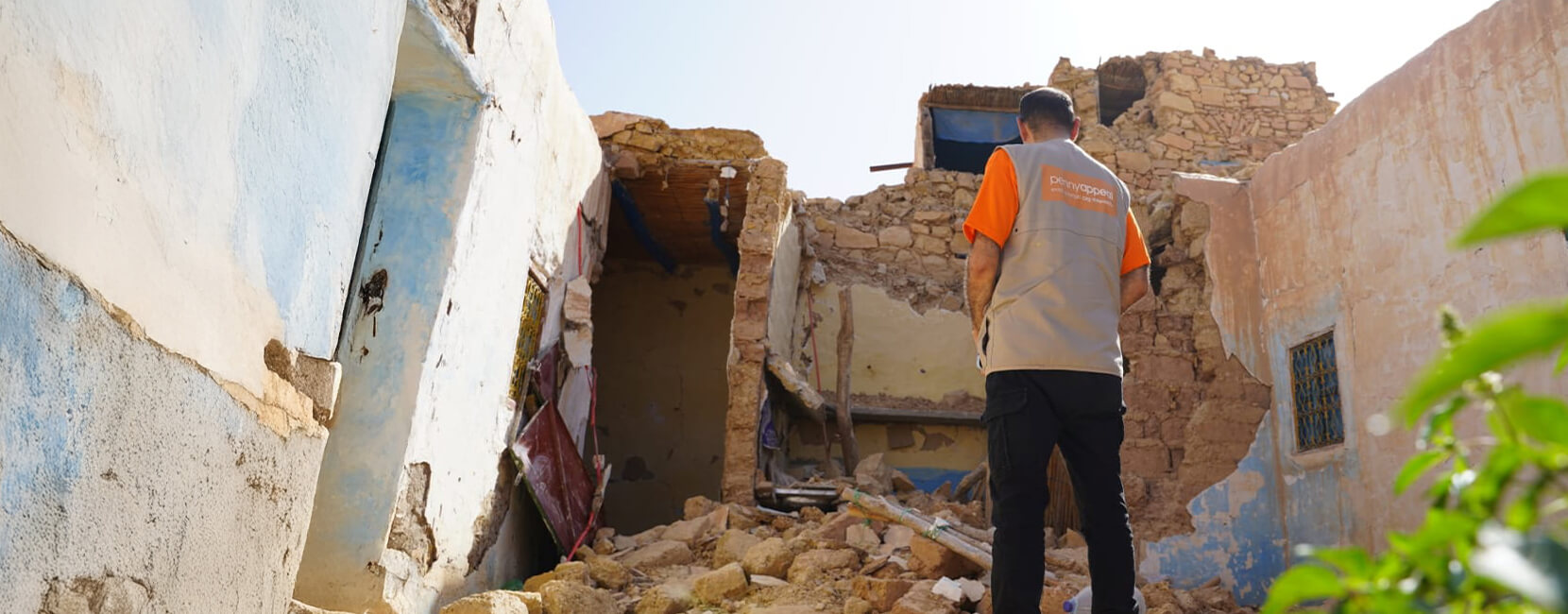 A Penny Appeal aid worker stands among the rubble of a collapsed building, assessing damage in a disaster-stricken area under clear skies