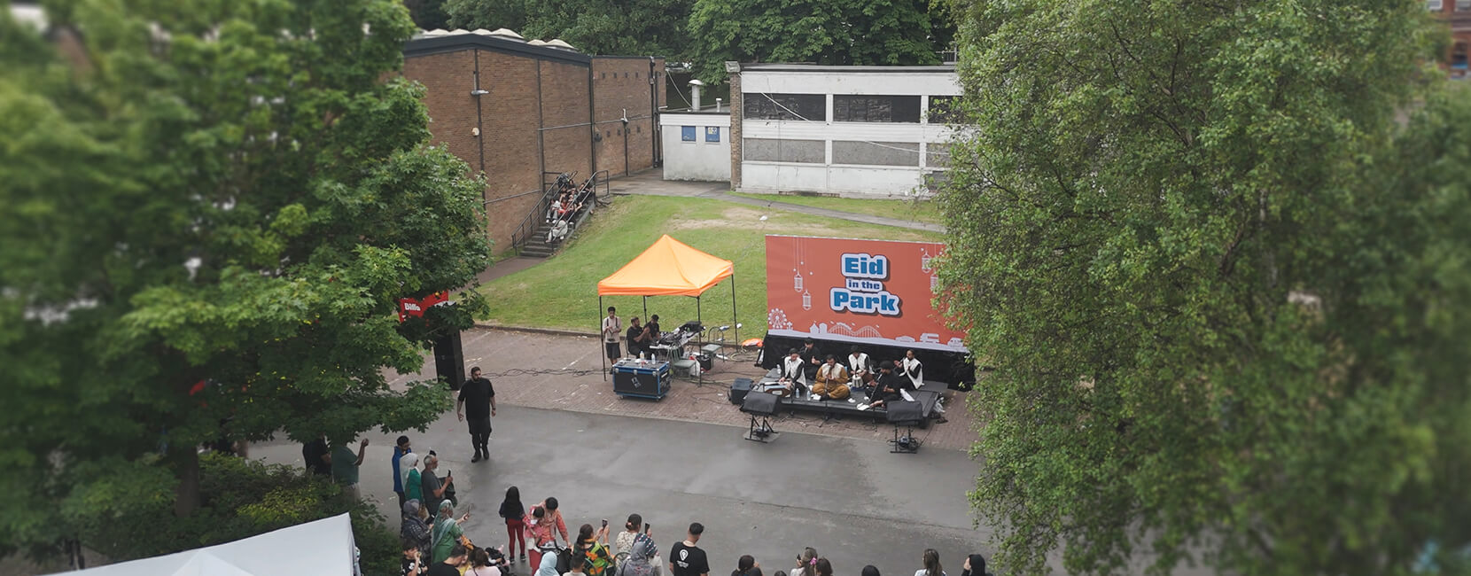 A community event organized by Penny Appeal called 'Eid in the Park,'' with people gathered around a stage featuring live performances under an orange canopy, surrounded by trees and urban buildings.
